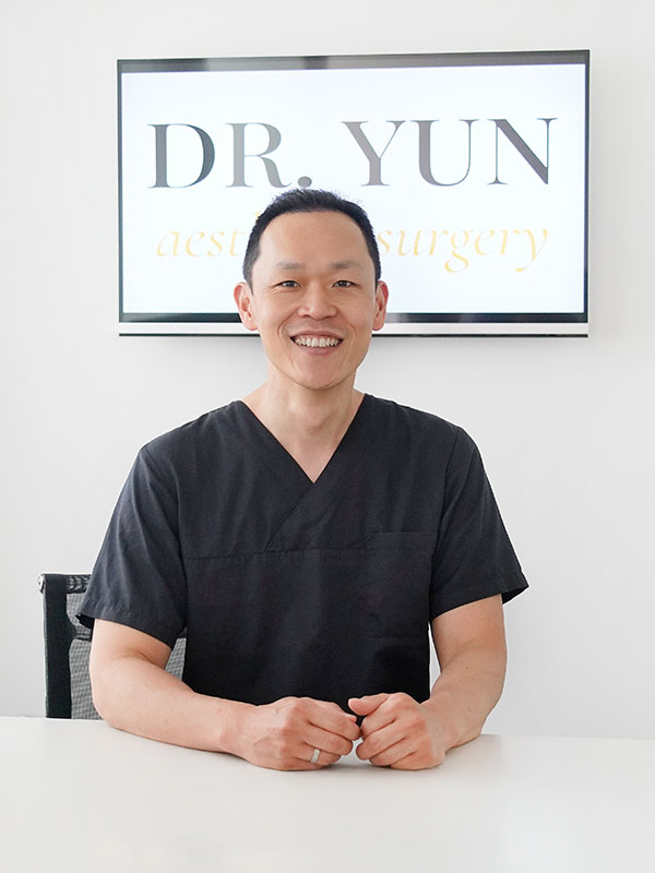 Dr. Yun sitzt l&auml;chelnd am Schreibtisch, hinter Ihm das Praxislogo.