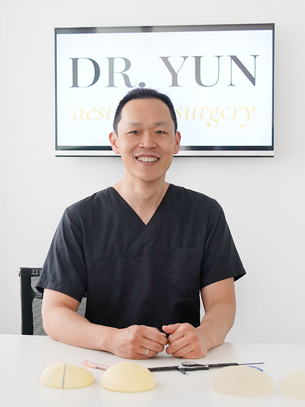 Dr. Yun sitzt l&auml;chelnd am Schreibtisch, hinter Ihm das Praxislogo.