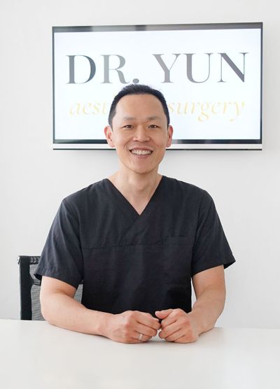 Dr. Yun sitzt lächelnd am Schreibtisch, hinter Ihm das Praxislogo.