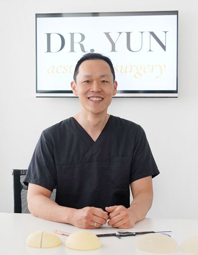 Dr. Yun sitzt lächelnd am Schreibtisch, hinter Ihm das Praxislogo.