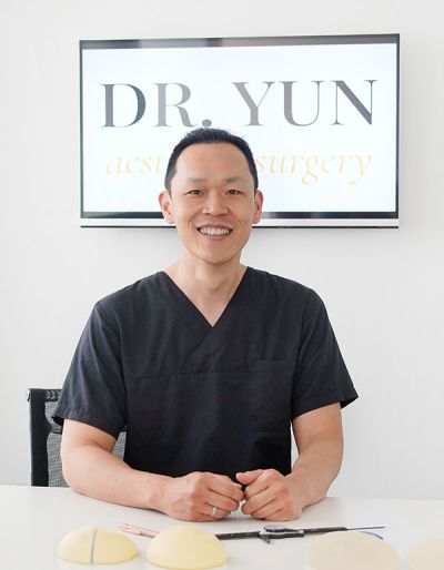 Dr. Yun sitzt lächelnd am Schreibtisch, hinter Ihm das Praxislogo.