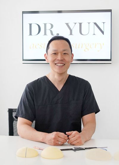 Dr. Yun sitzt lächelnd am Schreibtisch, hinter Ihm das Praxislogo.