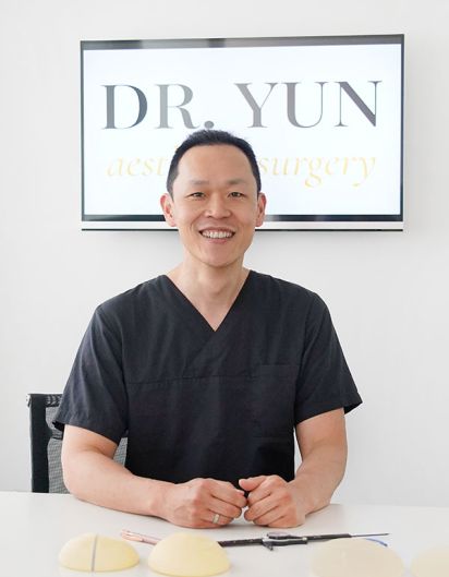 Dr. Yun sitzt lächelnd am Schreibtisch, hinter Ihm das Praxislogo.